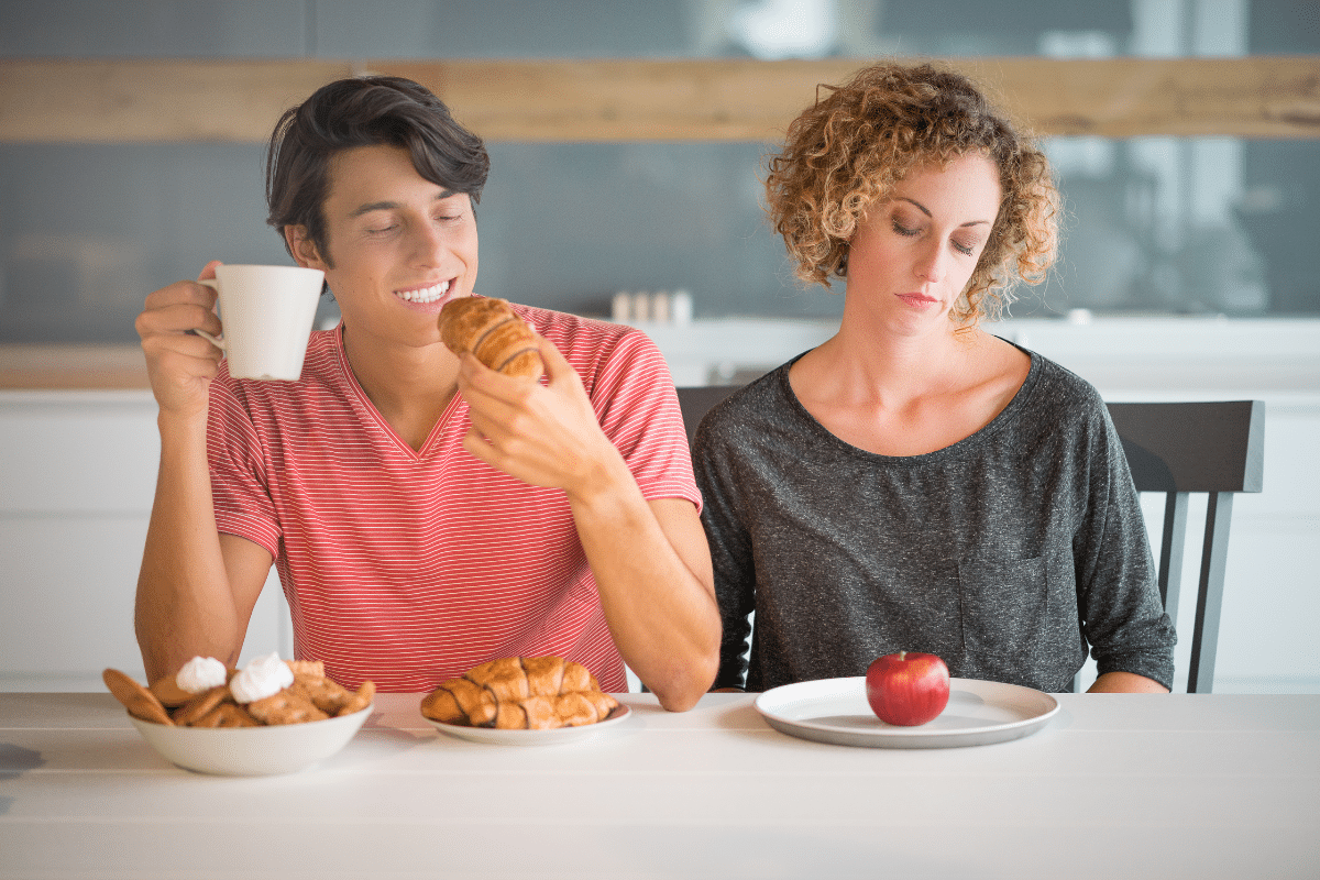 man eating lots of pasries whilst a woman has a plate with an apple on it.  she doesn't look happy